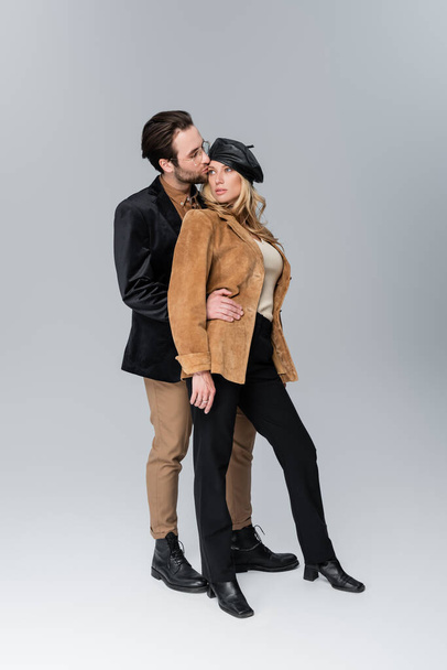 full length of bearded man in sunglasses kissing blonde woman in stylish beret on grey - Photo, Image