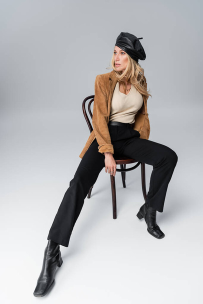full length of blonde woman in stylish leather beret and beige blazer posing while sitting on chair on grey - Fotografie, Obrázek