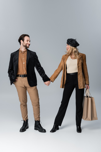 happy man holding hands with blonde woman in stylish beret standing with shopping bags on grey - Foto, afbeelding