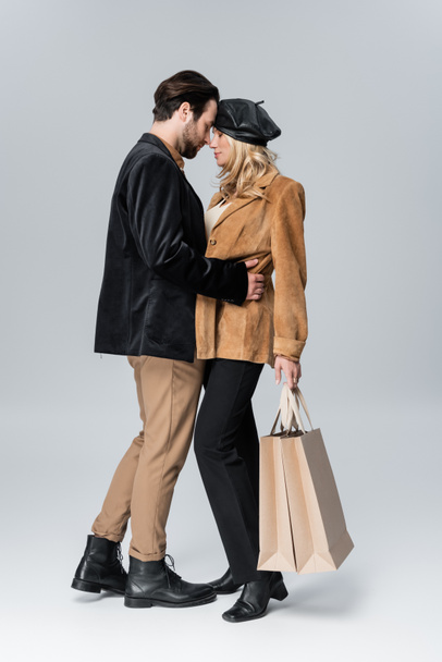 side view of bearded man hugging blonde girlfriend in stylish beret holding shopping bags on grey - Foto, Imagen