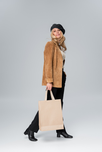 full length of blonde model in stylish leather beret and beige blazer holding shopping bag on grey - Fotó, kép