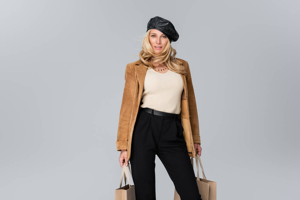 Улыбающаяся женщина в стильном кожаном берете и бежевом пиджаке держит сумки, изолированные на сером - Фото, изображение