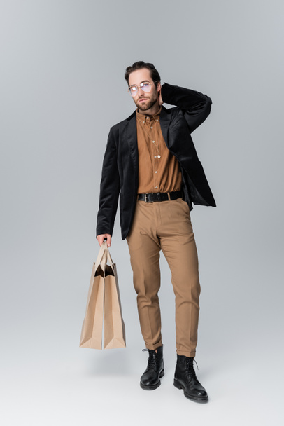 full length of bearded man in sunglasses and trendy autumnal outfit posing with shopping bags on grey - Foto, Imagem