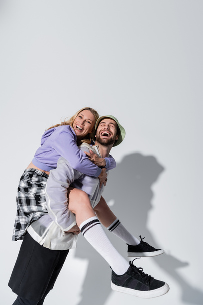 amazed man in panama hat piggybacking joyful woman in longs socks and sneakers on grey - Fotoğraf, Görsel
