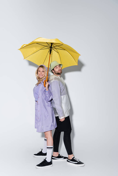 full length of happy woman in tartan skirt standing with bearded man in panama hat under yellow umbrella on grey - Photo, image