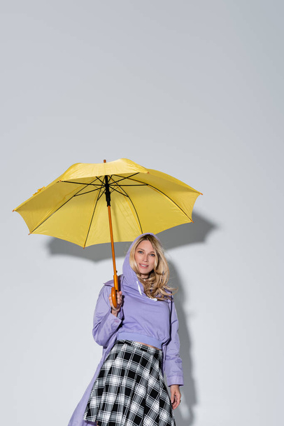 happy woman in tartan skirt smiling standing under yellow umbrella on grey - Фото, изображение