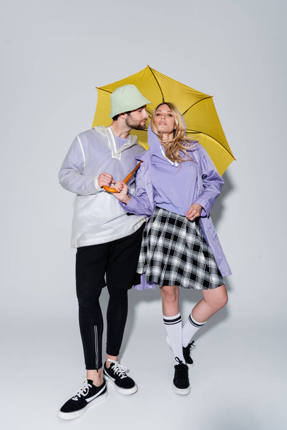 full length of woman in tartan skirt and longs socks posing with man in panama hat under yellow umbrella on grey - 写真・画像