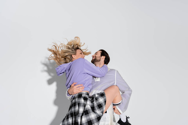 cheerful man holding in arms smiling blonde woman in tartan skirt on grey - Foto, afbeelding