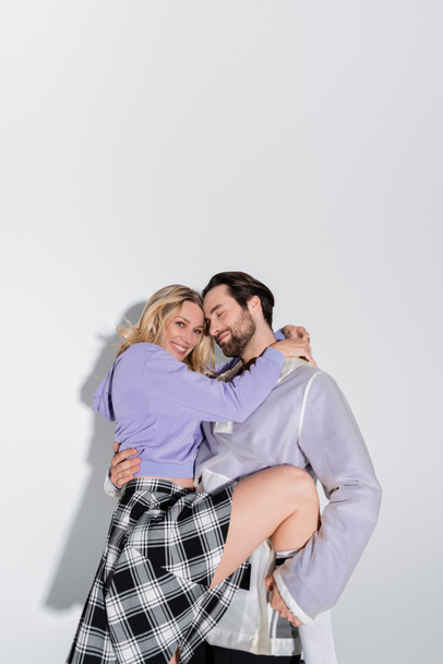 cheerful man holding in arms smiling blonde woman in tartan skirt on grey - 写真・画像