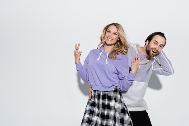 excited man near cheerful woman in purple sweatshirt showing peace sign on grey - Foto, Imagem