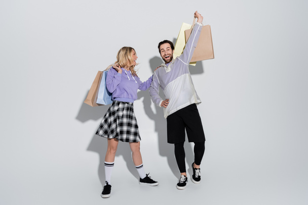 full length of excited man and cheerful woman in tartan skirt holding shopping bags on grey - Foto, Imagen