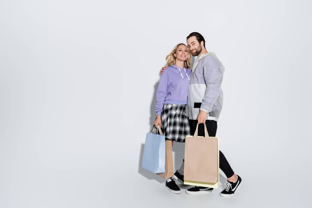 full length of happy man and blonde woman in tartan skirt holding shopping bags on grey - Foto, imagen