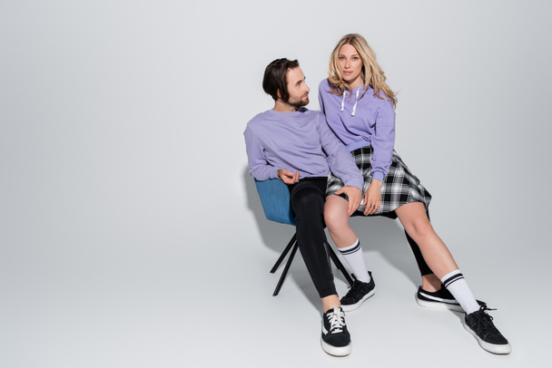 full length of cheerful man and blonde woman in tartan skirt sitting on blue armchair on grey - Foto, immagini