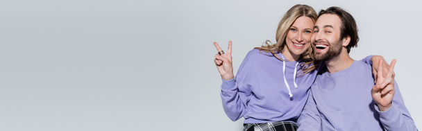 happy couple in purple sweatshirts showing peace sign and smiling isolated on grey, banner - Foto, Imagem