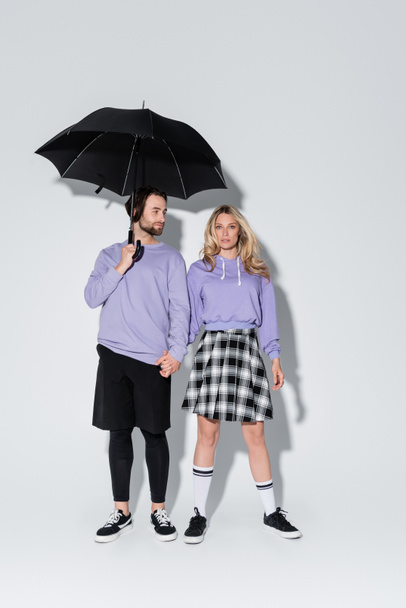full length of stylish couple in purple sweatshirts standing under black umbrella on grey - Fotografie, Obrázek