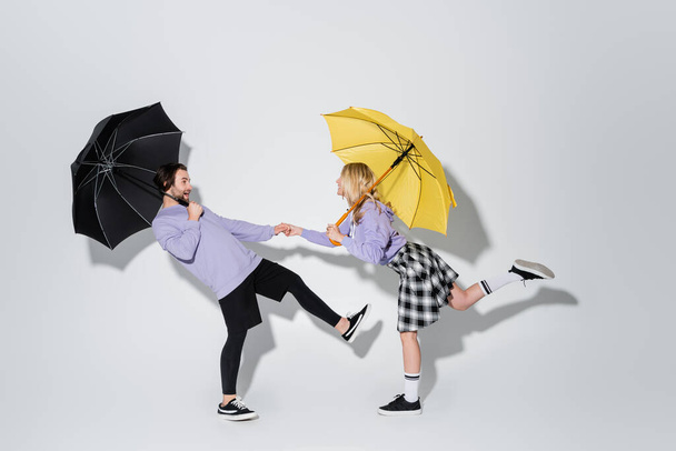 full length of cheerful couple in purple sweatshirts holding hands while standing under umbrellas on grey - Фото, изображение