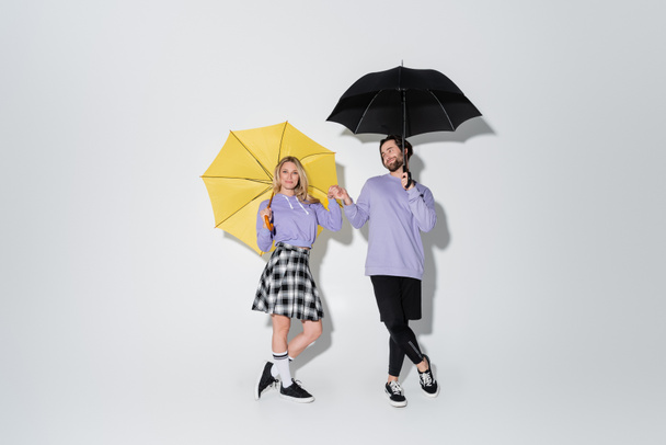 full length of pleased couple in purple sweatshirts holding hands while standing under umbrellas on grey - 写真・画像