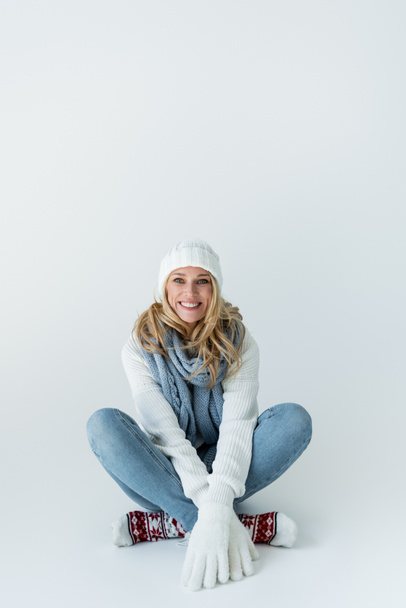 happy blonde woman in winter hat and knitted scarf looking at camera while sitting on grey - Foto, Bild