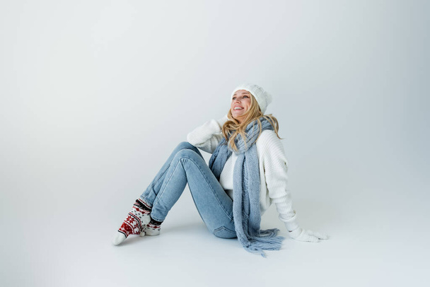 joyful blonde woman in white knitted hat and winter outfit sitting on grey - 写真・画像