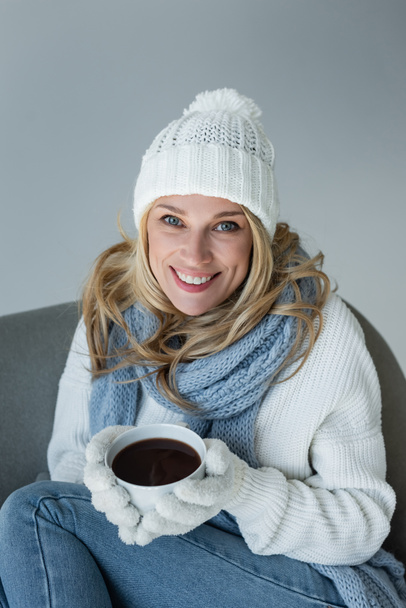 happy blonde woman in winter outfit sitting in comfortable armchair and holding cup of coffee isolated on grey - Foto, Bild