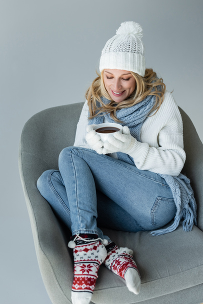 cheerful blonde woman in winter outfit sitting in comfortable armchair and holding cup of coffee isolated on grey - Foto, immagini