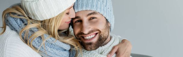 blonde woman in knitted hat hugging smiling boyfriend in winter outfit isolated on grey, banner - Photo, image