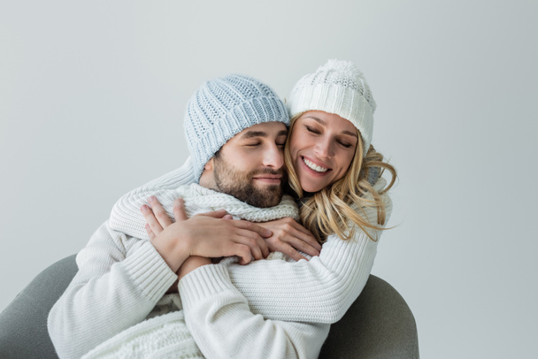 cheerful woman in knitted hat hugging joyful boyfriend in winter outfit isolated on grey - Photo, Image