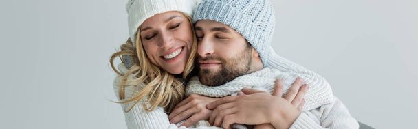 счастливая блондинка в вязаной шляпе, обнимающая улыбающегося бойфренда в зимнем наряде, изолированном на сером, баннере - Фото, изображение