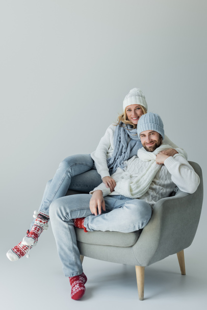 ευτυχισμένη ξανθιά γυναίκα σε πλεκτό καπέλο αγκαλιάζει με χαμογελαστό φίλο σε χειμερινό ντύσιμο, ενώ κάθεται σε πολυθρόνα σε γκρι - Φωτογραφία, εικόνα