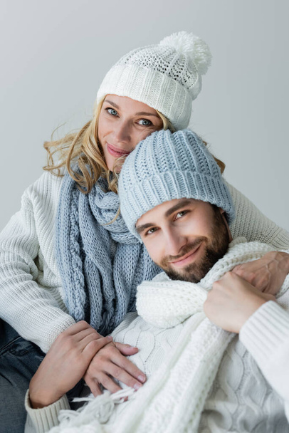 cheerful woman in knitted scarf hugging bearded boyfriend in winter hat isolated on grey - Fotoğraf, Görsel