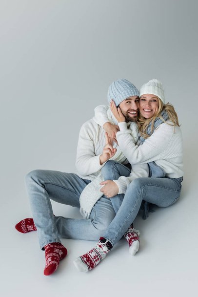 full length of happy blonde γυναίκα σε πλεκτό καπέλο αγκαλιάζει χαρούμενο φίλο σε χειμωνιάτικο ντύσιμο, ενώ κάθεται σε γκρι - Φωτογραφία, εικόνα
