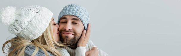happy blonde woman in knitted hat kissing cheek of smiling boyfriend isolated on grey, banner - Foto, imagen