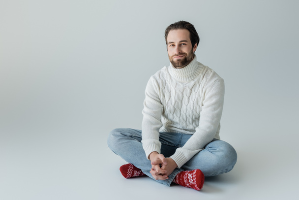full length of bearded man in jeans and knitted sweater sitting in red socks with ornament on grey  - Foto, imagen