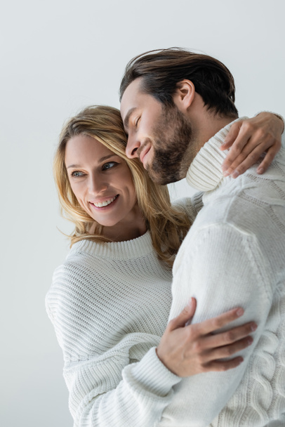 cheerful couple in knitted white sweaters and jeans smiling while hugging isolated on grey - Foto, imagen