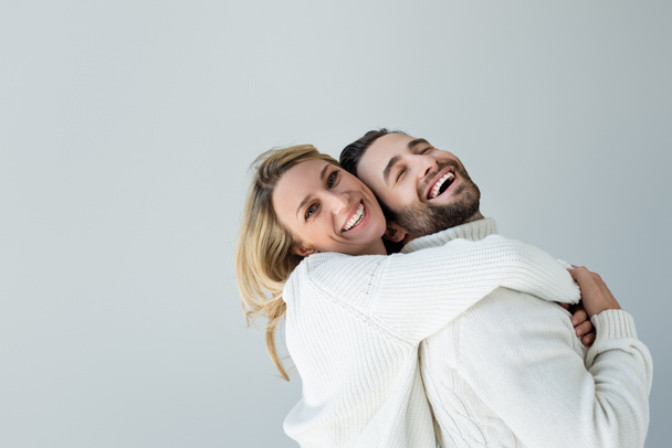 positive blonde woman in white knitted sweater hugging bearded man isolated on grey - Foto, imagen