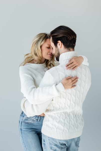 cheerful woman in white sweater hugging with boyfriend isolated on grey - Фото, изображение