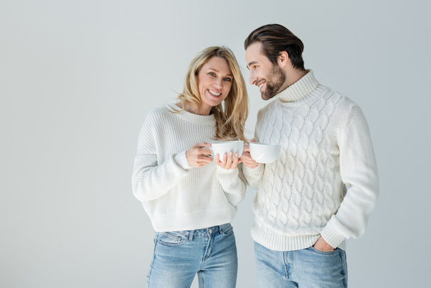 happy man and cheerful woman in white knitted sweaters holding cups of coffee isolated on grey - Фото, зображення