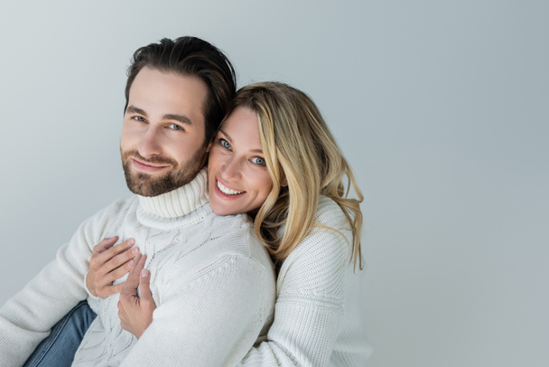 cheerful woman hugging bearded man in white sweater and looking at camera isolated on grey - 写真・画像