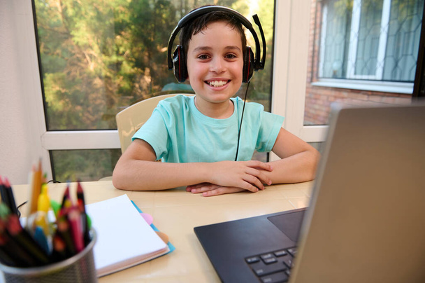 Portrait of a confident smart schoolboy wearing wireless headphones, smiling at camera, sitting at laptop, enjoying learning online foreign language courses. Homeschooling. Child development - Photo, Image