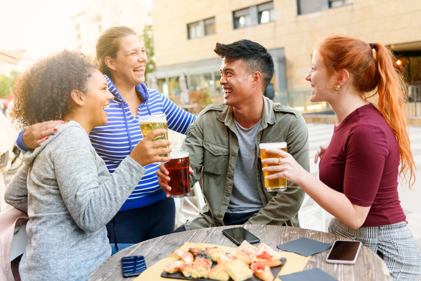 Счастливая женщина смеется и обнимает разнообразных друзей с пивом сидя за столом паба с закусками и смартфонами на городской улице - Фото, изображение