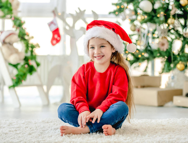 Little girl child wearing Santa hat in Christmas time sitting and smiling at decorated New Year home with festive tree. Cute kid in Xmas time - Fotografie, Obrázek