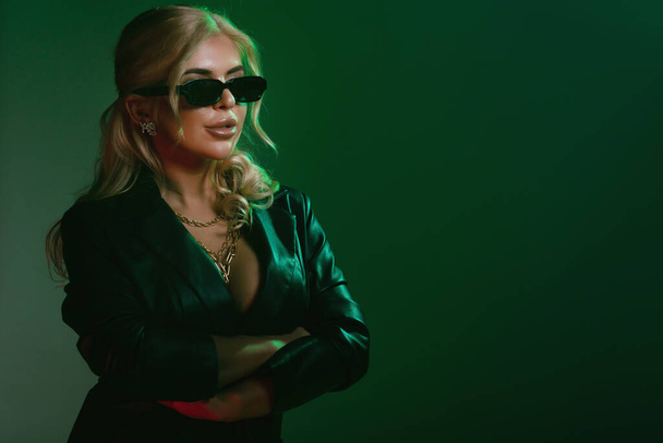 Studio fashion shot of the beautiful rock woman dressed in black leather jacket and black body lingerie, sunglasses on the face. Seductive lady in green light on a dark green background - Photo, Image