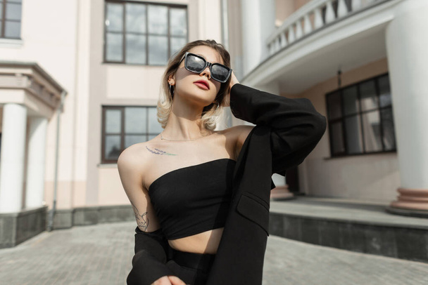Trendy cool vogue elegant woman with stylish modern sunglasses in black top with fashion blazer walks in the city near a vintage building - Zdjęcie, obraz
