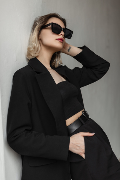 Trendy fashionable young woman model with short hair and sunglasses in fashionable business elegant black clothes stands near a white wall on the street - Photo, image