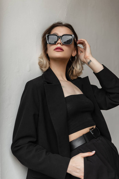 Cool elegant fashion young woman with stylish eyewear in black fashionable business outfit with top and blazer poses near a white wall on the street - Fotó, kép