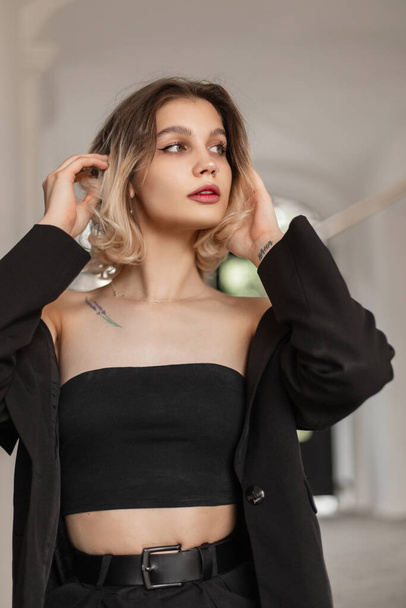 Fashionable beautiful young woman with cute face and hairstyle in black stylish clothes with top and blazer walks and poses on the street - Φωτογραφία, εικόνα