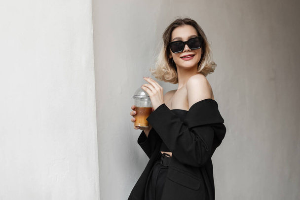 Щаслива молода елегантна модна усміхнена модель жінки в чорному стильному одязі з верхнім, чорним модним блейзером і штанами п'є холодну каву і ходить на вулиці біля білої стіни
 - Фото, зображення