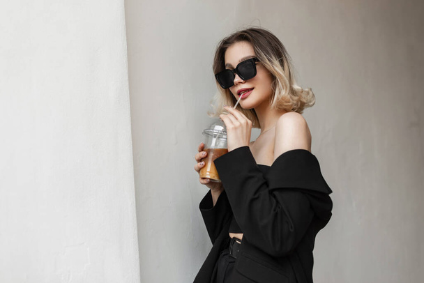 Κομψή όμορφη νεαρή όμορφη γυναίκα με μοντέρνα δροσερά γυαλιά ηλίου σε κομψά μαύρα ρούχα με κορυφή και σακάκι πίνοντας κρύο καφέ με χυμό και περπατώντας στο δρόμο - Φωτογραφία, εικόνα