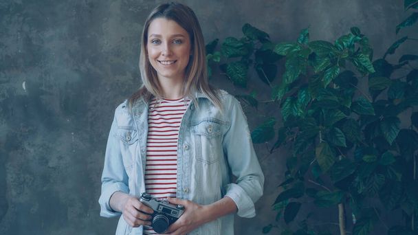 Portrait of pretty young blonde standing in loft apartment, smiling happily, posing and looking at camera. She is wearing fashionable denim jacket and top and holding camera. - Photo, Image