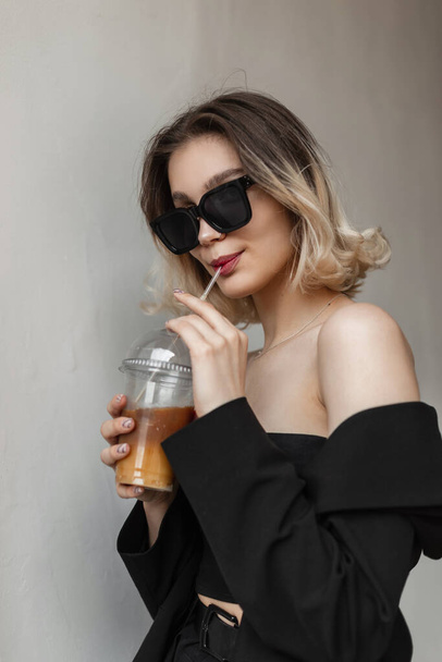 Fashionable glamour young elegant woman with black sunglasses in black business outfit drinks coffee and walks on the street. Urban girl lifestyle - Foto, imagen